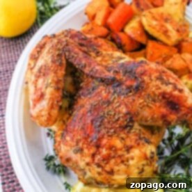 Half roasted chicken with herbs is elegantly served alongside fresh vegetables and lemon slices on a pristine white platter.