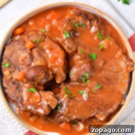 A plate of braised beef shin in rich tomato sauce with parsley garnish, resting on a napkin with handles in the background.