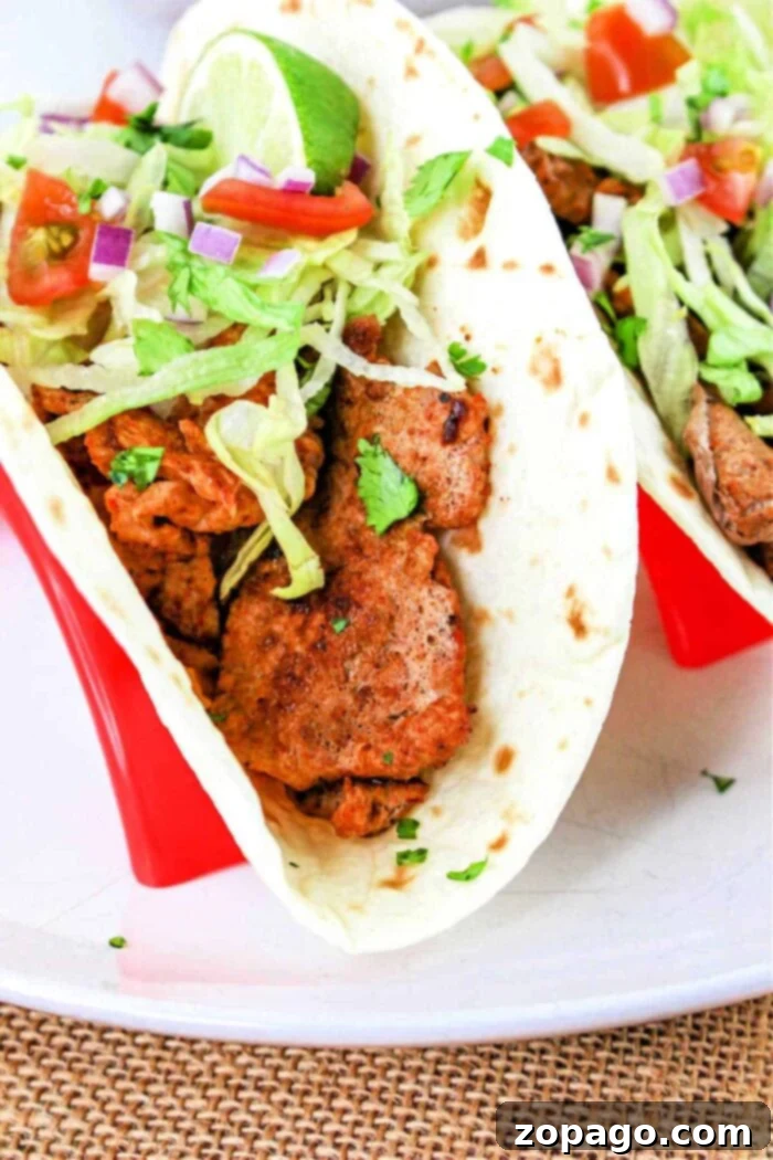 Succulent Pork Tenderloin Tacos 2 A close-up of a Pork Tenderloin Taco filled with seasoned meat, shredded lettuce, chopped tomatoes, diced red onions, and cilantro, all inside a soft flour tortilla. The taco is placed in a red taco holder. A lime wedge is also visible on top of the filling.