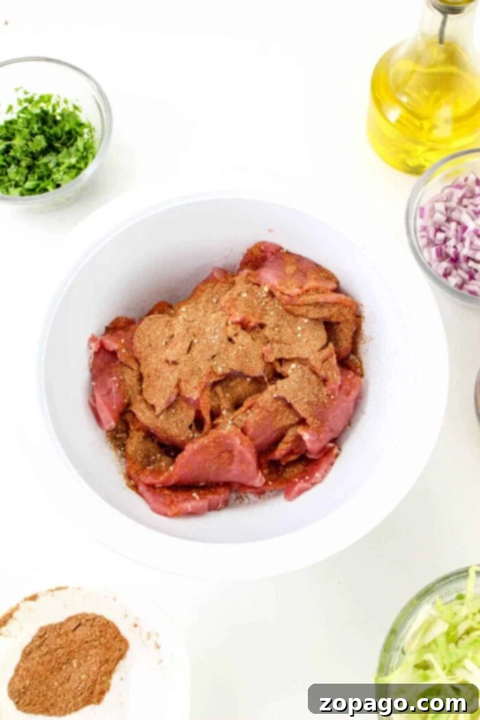 Succulent Pork Tenderloin Tacos 5 A white bowl contains raw pork pieces seasoned with a brown spice mix, surrounded by bowls of chopped herbs, diced red onions, shredded lettuce, a small bowl of additional spices resembling the ones used in pork tenderloin tacos, and a bottle of olive oil on a white background.