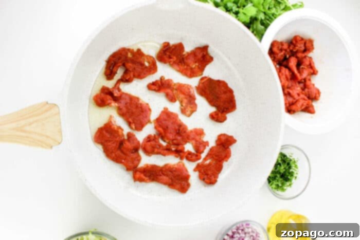 Succulent Pork Tenderloin Tacos 6 A white frying pan with raw marinated beef slices being cooked. Chopped cilantro, a bowl of additional raw beef, chopped red onions, minced garlic, and a small container of olive oil are arranged around the pan. Some fresh cilantro is also visible, evoking the vibrant setup for Pork Tenderloin Tacos.