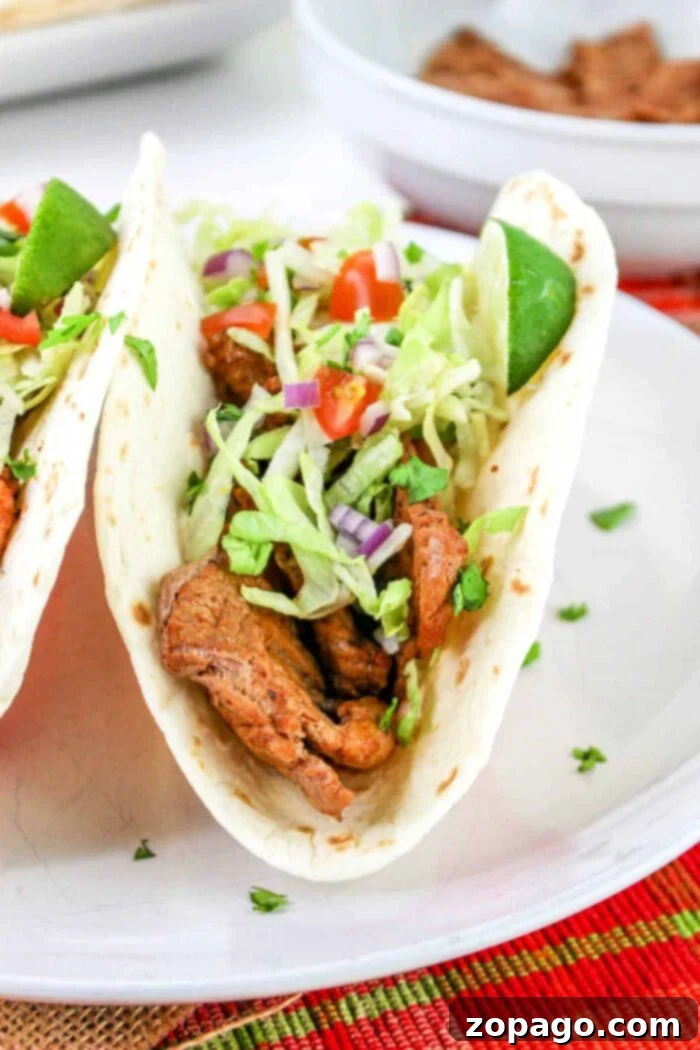 Succulent Pork Tenderloin Tacos 7 A close-up of a pork tenderloin taco filled with grilled pieces of meat, shredded lettuce, chopped tomatoes, and diced purple onions, garnished with a lime wedge. The taco is placed on a white plate with part of another taco visible in the background.