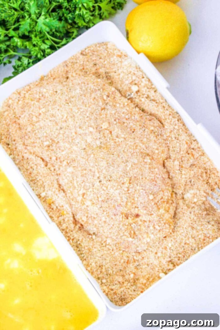 Perfectly Baked Chicken Cutlets 7 Breaded chicken breast cutlet on a tray, ready to answer the question of how long to bake chicken cutlets, with egg wash and parsley nearby.