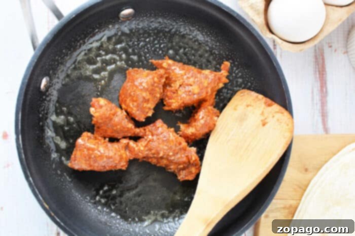 Spicy Chorizo Scramble 4 A black skillet on a counter, filled with sizzling pieces of chorizo, with a wooden spatula resting inside. Nearby, a carton of fresh eggs and a tortilla on a wooden cutting board await their turn, highlighting the essential steps in how to cook chorizo and eggs.