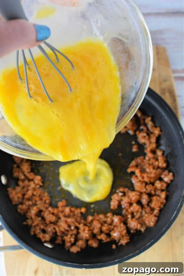 Spicy Chorizo Breakfast Scramble 5 A hand is seen pouring whisked eggs from a bowl into a frying pan that contains browned ground meat, possibly chorizo. The pan rests on a wooden surface. The person is holding a whisk in the bowl, which is slightly tilted. This demonstrates how to cook chorizo and eggs effortlessly.