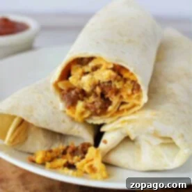 A white plate holds two burritos made with flour tortillas. One burrito is partially cut open, revealing a filling of scrambled eggs, sausage, and melted cheese. The scene offers a tempting glimpse into how to cook chorizo and eggs. Brown sauce in a small white bowl appears blurred in the background.