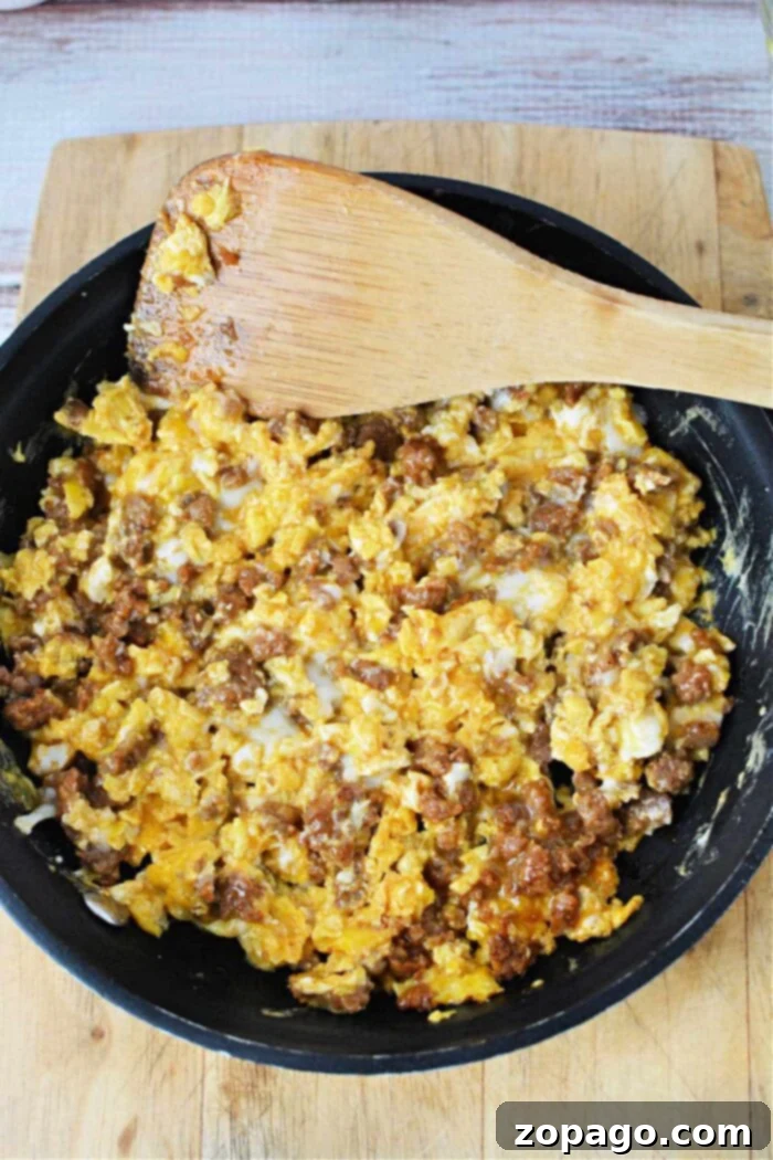 Spicy Chorizo Breakfast Scramble 7 A frying pan on a wooden cutting board contains scrambled eggs mixed with ground sausage. A wooden spatula rests on the edge of the pan, partially covered in the egg and sausage mixture—a perfect demonstration of how to cook chorizo and eggs.