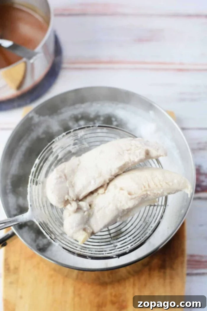 Chicken Tender Boiling Times 6 Two perfectly cooked chicken tenders are carefully lifted from a pot of simmering water using a wire skimmer, poised to be placed on a rustic wooden cutting board. This image captures the moment of doneness, showing how long to boil chicken tenders for ideal tenderness.