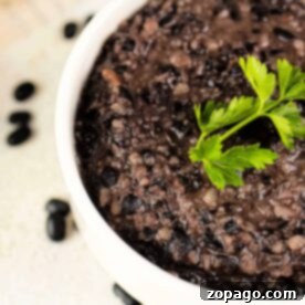 Pantry Staple Refried Black Beans 7 A delightful bowl of refried black beans recipe, topped with a fresh sprig of parsley, sits amid scattered black beans on the surface, inviting you to savor every bite.