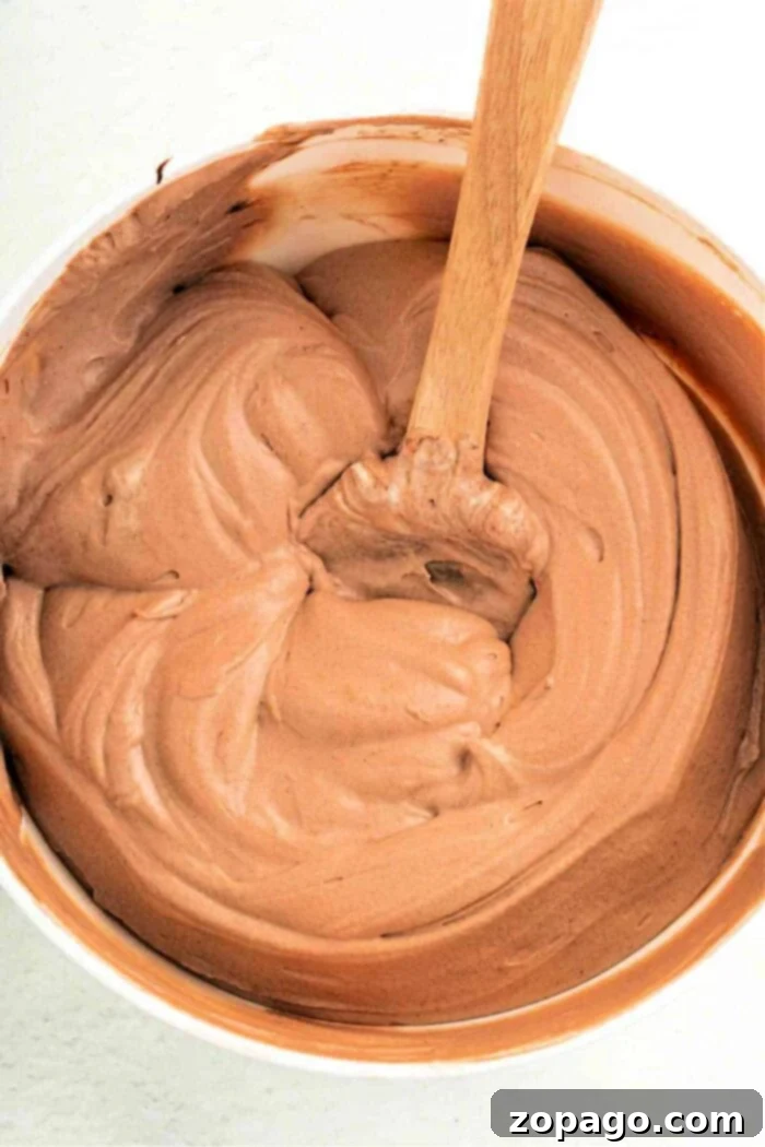 Dreamy Cool Whip Pudding Frosting 2 A bowl of creamy chocolate pudding frosting, enhanced with a dollop of Cool Whip, is being expertly stirred with a wooden spoon.