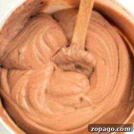 A bowl of creamy chocolate pudding frosting, enhanced with a dollop of Cool Whip, is being expertly stirred with a wooden spoon.