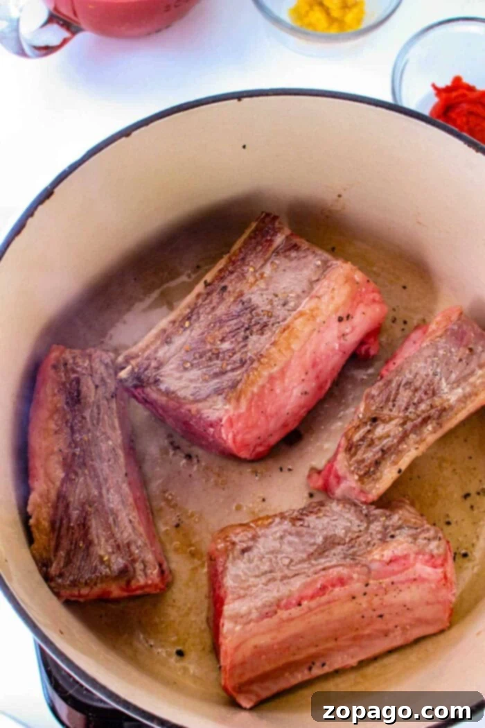 Slow-Cooked Short Rib Tacos 4 Four pieces of browned short rib meat are cooking in a large pot, perfect for tacos. The meat has a seared exterior. In the background, there are small bowls with ingredients, including a bright red and yellow substance. The pot and ingredients are set on a white surface.