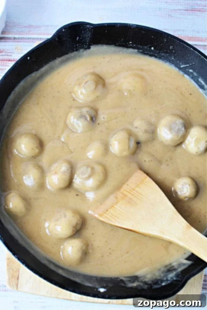 A skillet filled with succulent mushrooms smothered in a creamy brown sauce enriched with the flavors of cream of mushroom gravy, skillfully stirred with a wooden spatula.