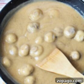 A skillet filled with succulent mushrooms smothered in a creamy brown sauce enriched with the flavors of cream of mushroom gravy, skillfully stirred with a wooden spatula.