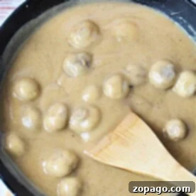 A skillet filled with succulent mushrooms smothered in a creamy brown sauce enriched with the flavors of cream of mushroom gravy, skillfully stirred with a wooden spatula.
