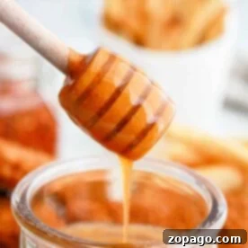 A honey dipper drizzling hot honey sauce into a glass jar