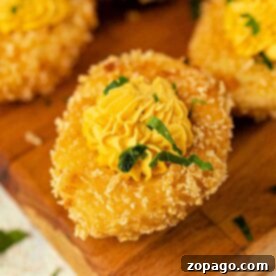 A crispy breaded deep-fried deviled eggs, topped with a yellow piped sauce and garnished with green herbs on a wooden board.