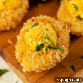 A crispy breaded deep-fried deviled eggs, topped with a yellow piped sauce and garnished with green herbs on a wooden board.
