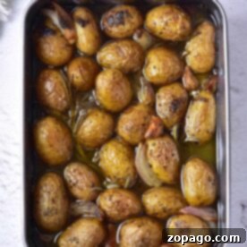 A baking tray of confit roasted baby potatoes with herbs and garlic cloves rests on a white surface.