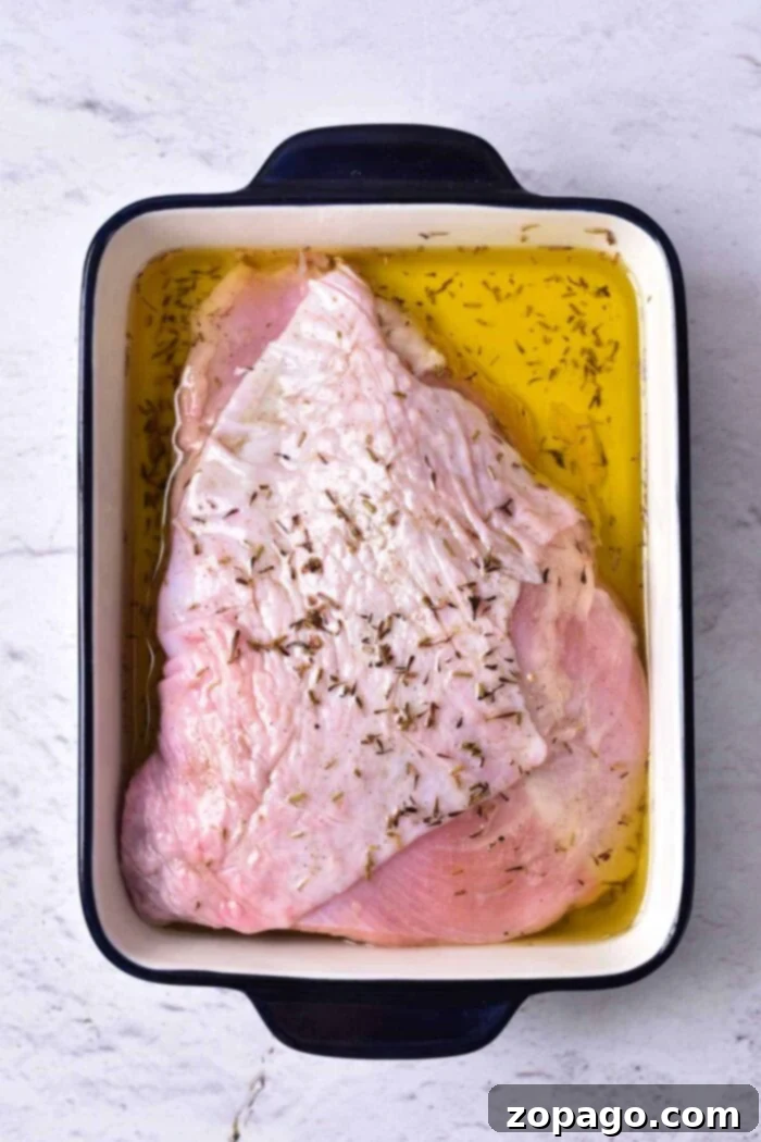 Slow Cooked Turkey Breast Confit 4 A raw turkey breast marinating in oil and seasonings, destined for a delicious turkey confit, sits in a rectangular white and black roasting pan on a light-colored surface.
