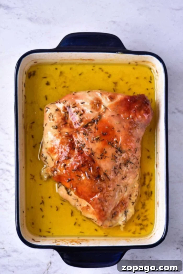 Slow Cooked Turkey Breast Confit 5 A cooked piece of turkey breast, prepared as Turkey Confit, is shown in a rectangular blue and white baking dish filled with golden liquid, likely broth or melted butter. The turkey is seasoned with herbs. The dish is placed on a light-textured surface.