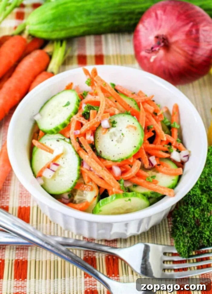 Refreshing Cucumber Carrot Salad 2 A white bowl filled with a Carrot and Cucumber Salad, featuring sliced cucumbers, julienned carrots, and chopped red onions, garnished with herbs. The bowl sits on a woven mat, surrounded by whole carrots, a cucumber, a red onion, and parsley. A fork and knife rest beside the bowl.