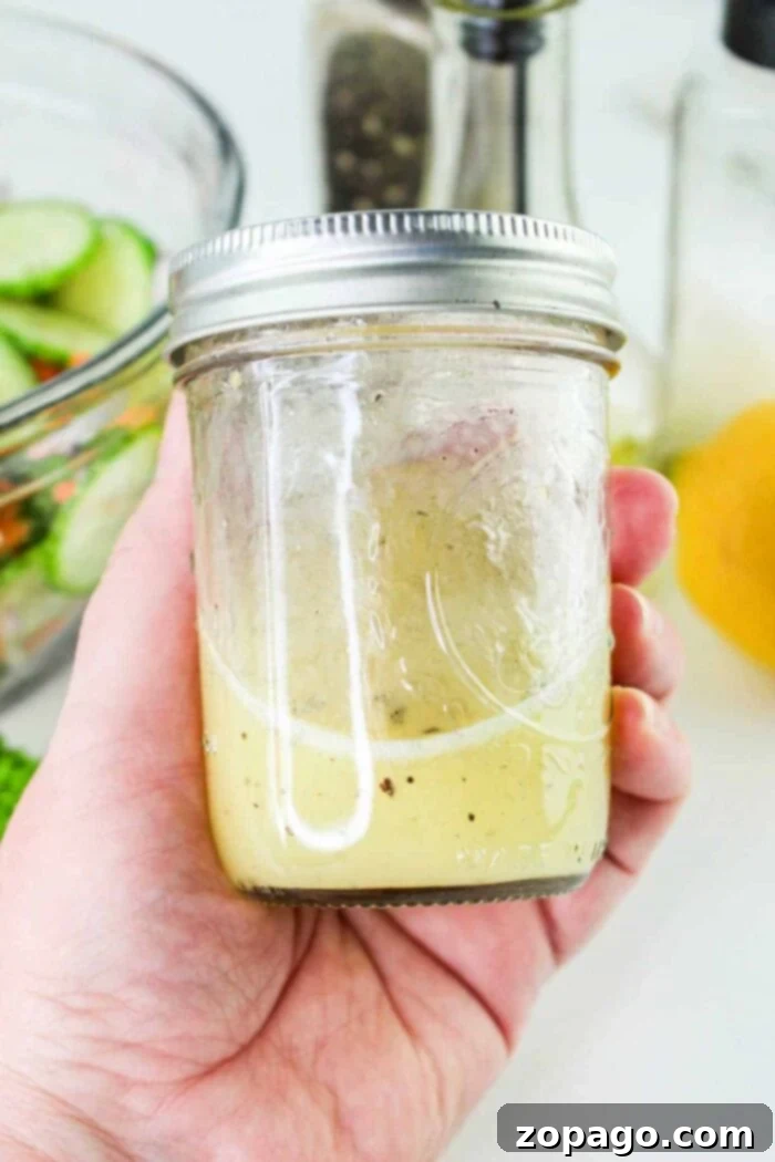 Refreshing Cucumber Carrot Salad 4 A hand is holding a glass jar filled halfway with light-colored salad dressing, perfect for a refreshing Carrot and Cucumber Salad. The jar has a metal screw-top lid. In the background, there is a bowl of sliced cucumbers, a pepper grinder, a bottle, and a lemon.