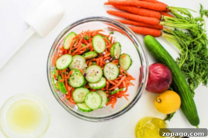 Refreshing Cucumber Carrot Salad 5 A glass bowl filled with a Carrot and Cucumber Salad, featuring sliced cucumbers, shredded carrots, and chopped herbs. Surrounding the bowl are whole carrots with greens, a cucumber, a red onion, a lemon, a small bowl of dressing, a bottle of oil, and a white kitchen tool.