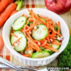 A white bowl filled with a Carrot and Cucumber Salad, featuring sliced cucumbers, julienned carrots, and chopped red onions, garnished with herbs. The bowl sits on a woven mat, surrounded by whole carrots, a cucumber, a red onion, and parsley. A fork and knife rest beside the bowl.