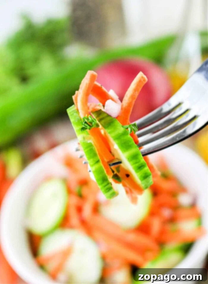 Refreshing Cucumber Carrot Salad 7 A fork holds a bite-sized portion of Carrot and Cucumber Salad, featuring cucumber slices, carrot strips, and chopped red onions. In the background, there is a bowl of similar salad ingredients and some vegetables.
