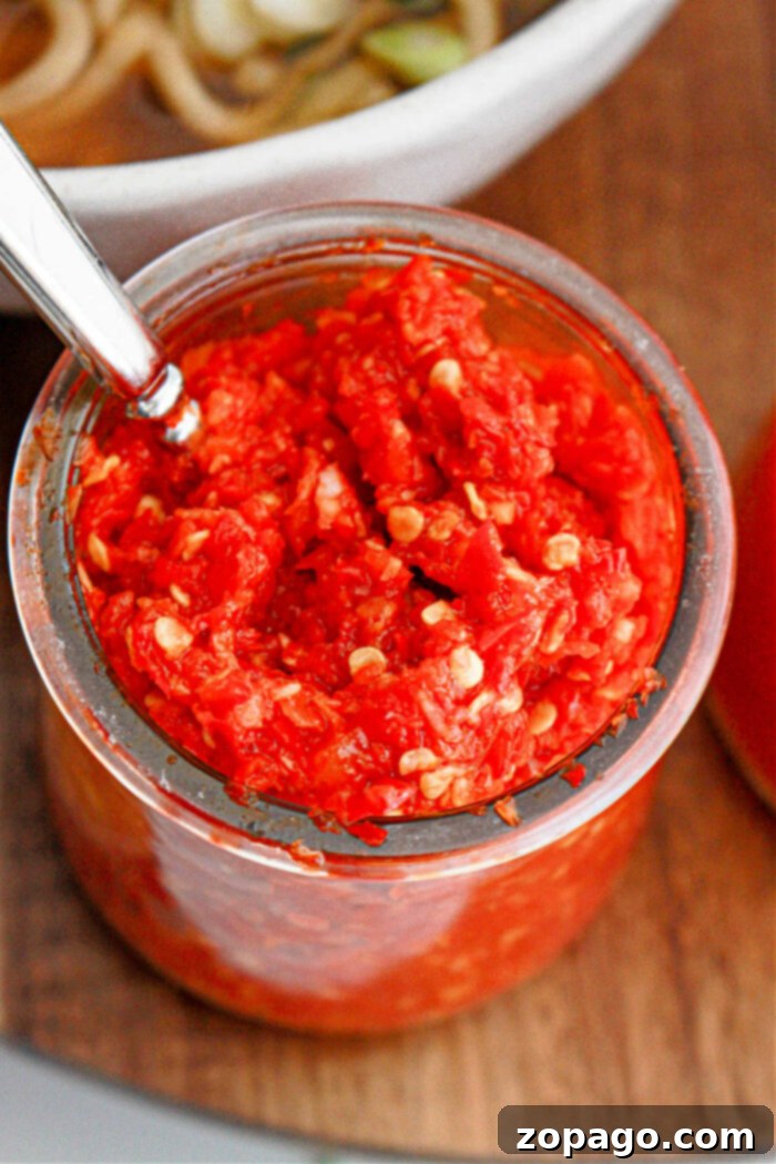 Fiery Garlic Chili Paste 6 A clear glass jar filled with chunky garlic chili paste, showing visible seeds and a metal spoon inserted into the jar. It sits on a wooden surface with part of a white bowl containing food visible in the background.