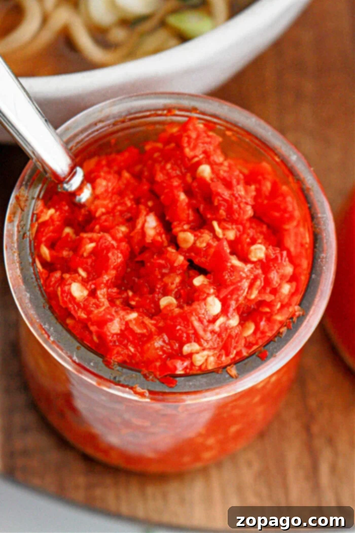 Homemade Fiery Chili Garlic Paste 6 A clear glass jar filled with chunky, bright red garlic chili paste, showcasing visible seeds and a metal spoon inserted into the jar. It rests on a rustic wooden surface with a white bowl of food partially visible in the background, inviting a taste.