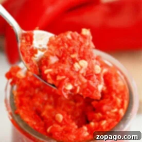 A close-up of a spoon holding a portion of vibrant red homemade garlic chili paste with visible seeds, positioned above a glass jar filled with the same rich mixture. In the soft-focus background, whole red peppers suggest the fresh ingredients used.