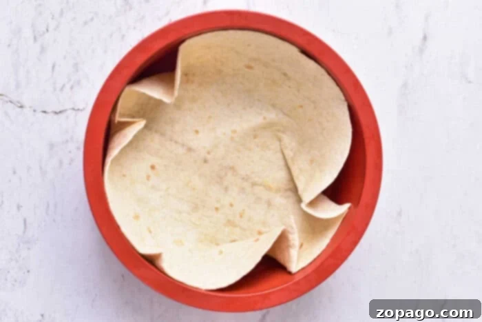 Wholesome Cottage Cheese Quiche 4 A single tortilla is arranged inside a red, round baking dish on a light-colored surface. The edges are slightly folded and overlapping, forming a semi-crumpled appearance, perfect for a delicious cottage cheese quiche. The background is textured with subtle cracks and lines.