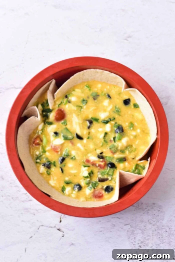 Creamy Cottage Cheese Quiche 6 A red bowl filled with a creamy yellow cottage cheese quiche mixture, abundant with chopped vegetables like olives, tomatoes, green peppers, and corn, all nestled within tortilla pieces. The background is a white, textured surface.
