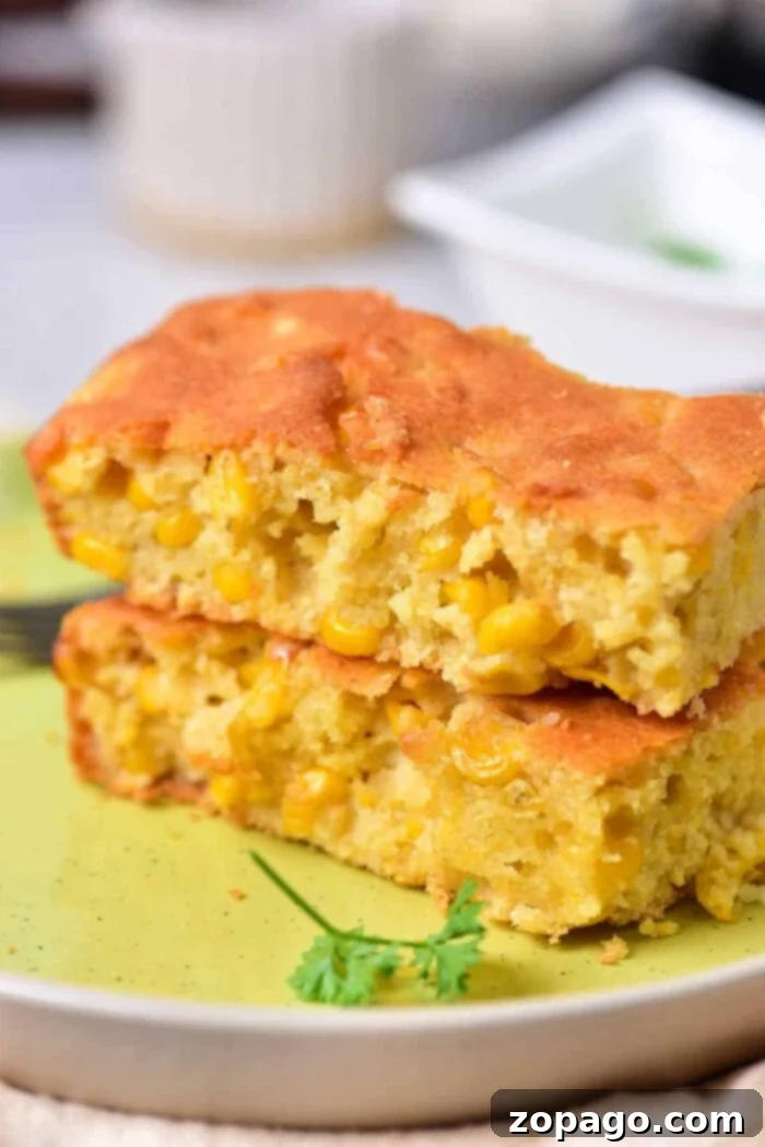 Homemade Creamy Corn Pudding 2 Two pieces of golden-brown corn pudding, studded with visible corn kernels, are stacked on a light green plate. A sprig of fresh parsley provides a vibrant garnish, with a fork partially visible in the background, inviting a taste of this traditional homemade corn pudding.