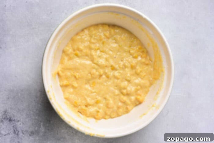 Homemade Creamy Corn Pudding 3 A white bowl brimming with a thick, yellowish batter, showcasing numerous visible corn kernels, rests on a light gray surface, perfectly prepared for baking into homemade Corn Pudding Without Jiffy Mix.