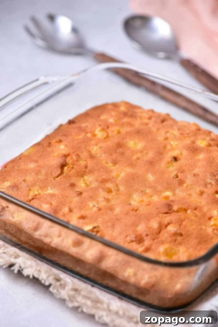 Homemade Creamy Corn Pudding 4 A square glass baking dish holds a freshly baked corn pudding with a golden-brown crust, reminiscent of homemade corn pudding without Jiffy mix. The dish is placed on a tan woven trivet, and two silver spoons are in the background on a light-colored surface.