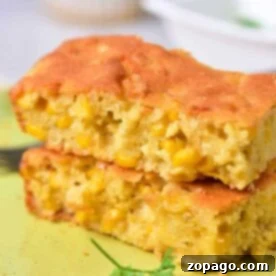 Two pieces of corn pudding with visible corn kernels stacked on a light green plate remind one of traditional Corn Pudding Without Jiffy Mix. A piece of parsley adorns the plate, and a fork is partially visible in the background.