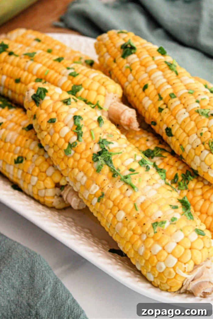 Perfect Slow Cooker Corn on the Cob 2 A close-up of several ears of Crockpot Corn on the Cob placed on a white rectangular dish. The corn is garnished with chopped fresh herbs and a sprinkle of seasoning. A green cloth is partially visible in the background.