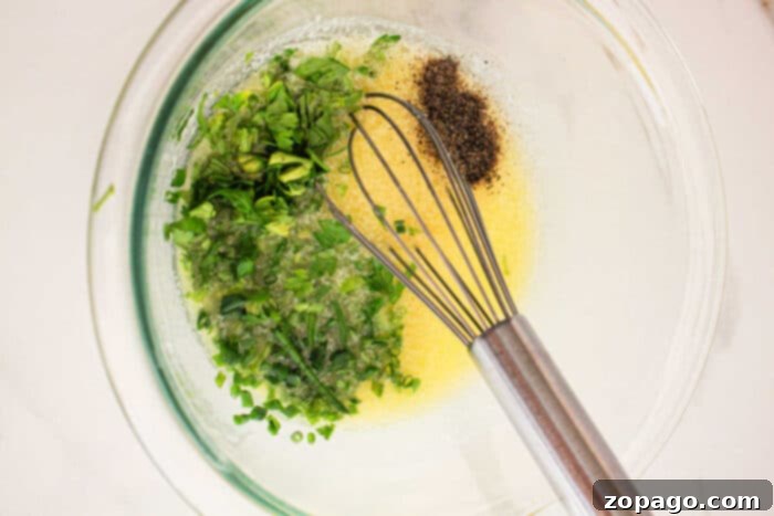 Perfect Slow Cooker Corn on the Cob 4 A glass mixing bowl containing a yellow liquid with finely chopped green herbs, ground black pepper, and a metal whisk, reminiscent of seasonings for Crockpot Corn on the Cob. The bowl is placed on a light-colored surface.