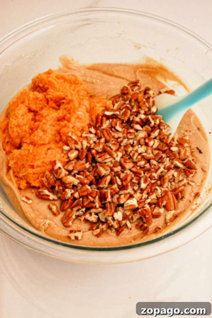 Spiced Sweet Potato Loaf 3 A glass bowl contains a mixture of ingredients being combined: creamy batter, orange sweet potato puree, and chopped pecans. A blue spatula is resting in the bowl, ready for mixing what will become delicious Sweet Potato Bread with Cake Mix.