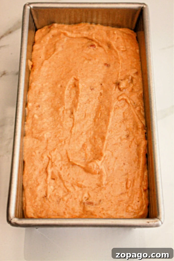 Spiced Sweet Potato Loaf 4 A metal loaf pan filled with unbaked sweet potato bread batter made with cake mix sits on a white surface. The batter appears smooth and evenly spread within the pan, ready for baking.