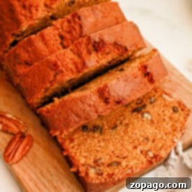 A loaf of sweet potato bread partially sliced on a wooden cutting board. The bread is golden brown with visible specks of nuts. Whole pecans and a cinnamon stick are placed beside the board on a white surface. A gray cloth is partially visible in the corner.