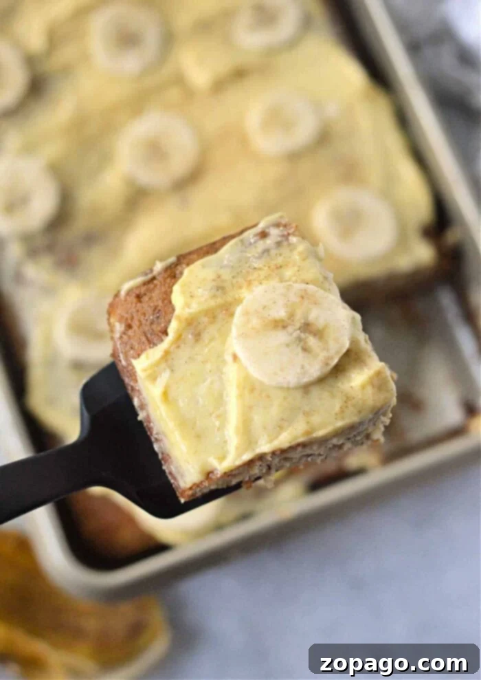 Cake Mix Banana Cake 2 A close-up of a spatula holding a slice of banana cake topped with creamy frosting and a banana slice. In the background, there is a baking tray containing more pieces of similarly topped treats from an easy banana cake recipe with cake mix.