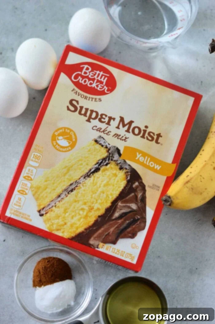 Cake Mix Banana Cake 3 A box of Betty Crocker Super Moist yellow cake mix is on a countertop. Around it are three eggs, a peeled banana, a cup of water, a small cup of oil, a bowl of cinnamon, and a bowl of baking soda—everything you need for an effortless banana cake recipe with cake mix.