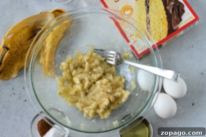 Cake Mix Banana Cake 4 A mixing bowl with mashed bananas and a fork sits on a countertop. Surrounding the bowl are two eggs, a box of cake mix, banana peels, a small bowl of cinnamon, and a can of baking ingredients—all the essentials for an easy banana cake recipe with cake mix.