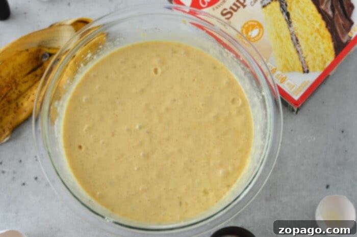 Cake Mix Banana Cake 5 A glass bowl filled with cake batter sits on a gray surface. Next to the bowl, there is a banana peel, a box featuring an enticing banana cake recipe with cake mix, and egg shells. The cake mix box shows a yellow cake with chocolate frosting.