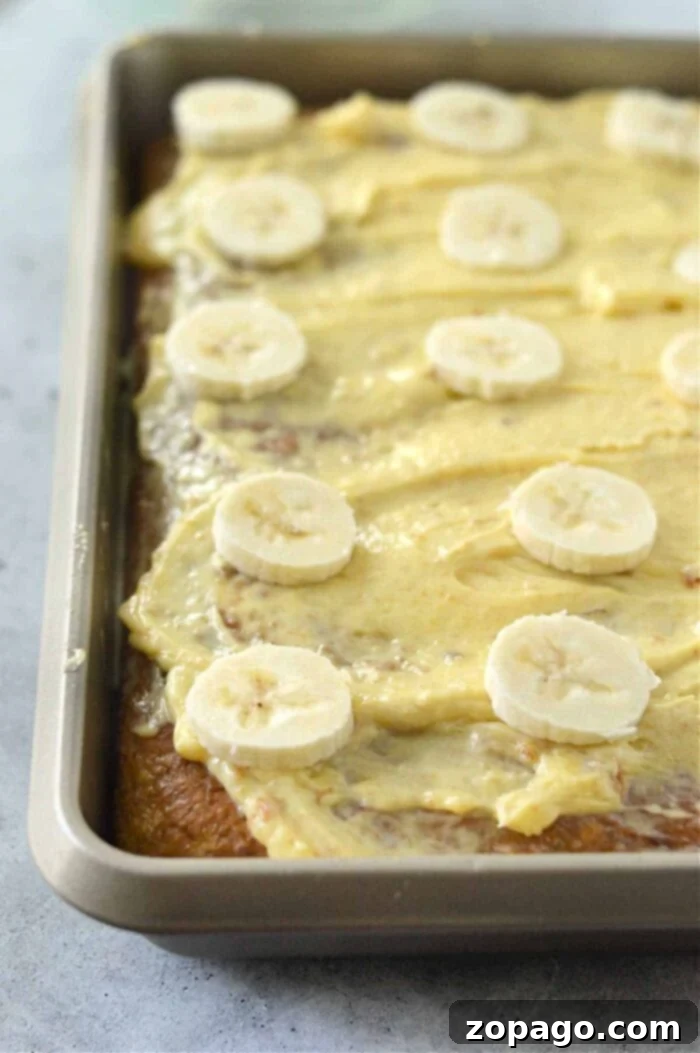 Cake Mix Banana Cake 7 A rectangular cake in a baking pan is topped with creamy frosting and sliced bananas. Using a banana cake recipe with cake mix, the frosting is spread evenly across the surface, and the banana slices are artfully arranged on top. The pan sits on a light gray countertop.
