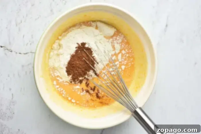 A mixing bowl filled with the beginnings of a pumpkin cake with yellow cake mix. It shows a vibrant yellow liquid base, likely beaten eggs and milk, combined with a heap of dry ingredients, including flour and cocoa powder. A silver whisk rests in the bowl on a light-colored surface, ready for blending.
